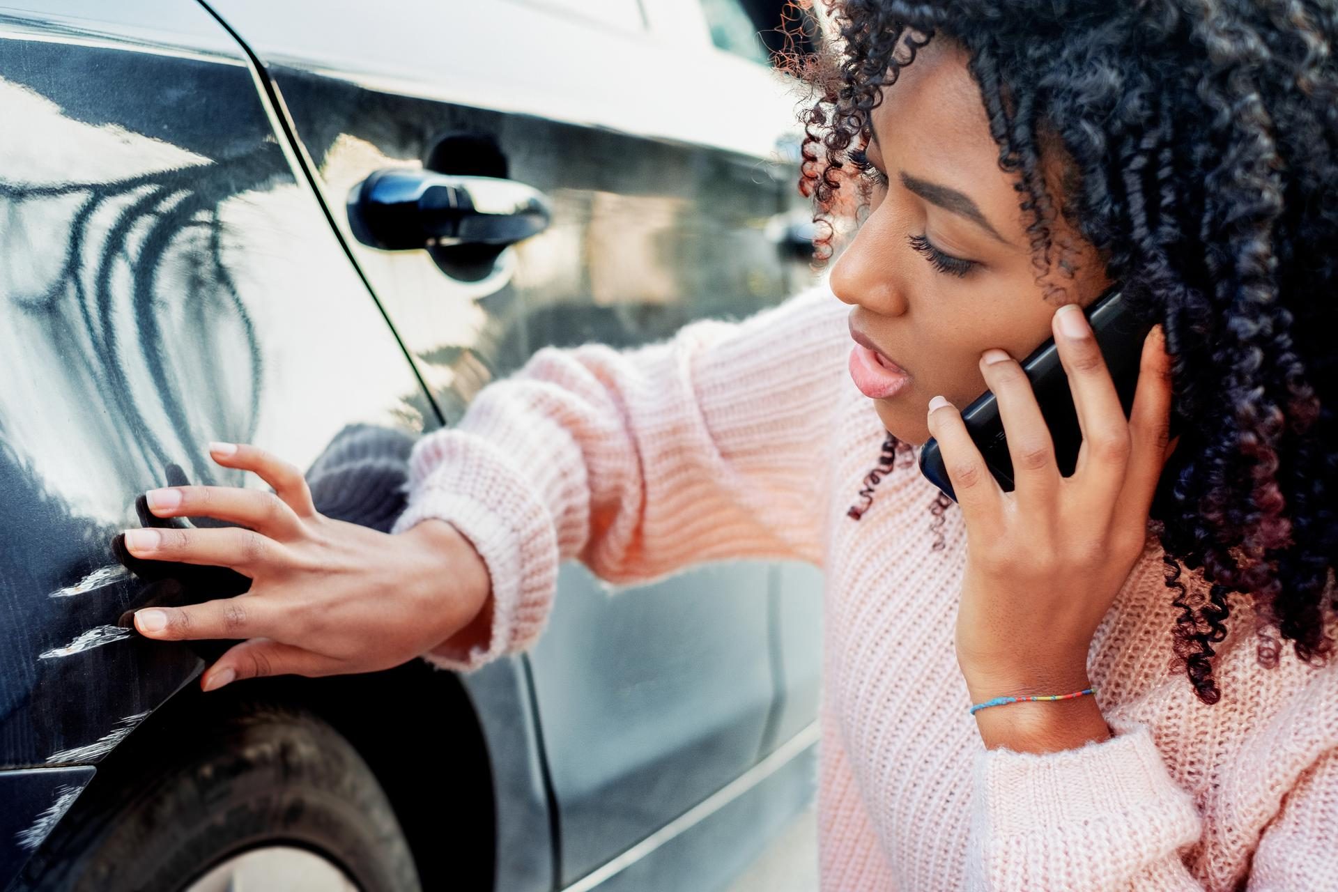 Zwarte jonge vrouw bekijkt autoschade en belt verzekeraar.